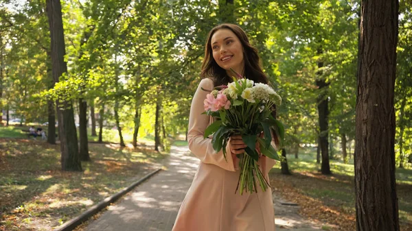 Cheerful brunette woman in dress holding bouquet in green park - foto de stock