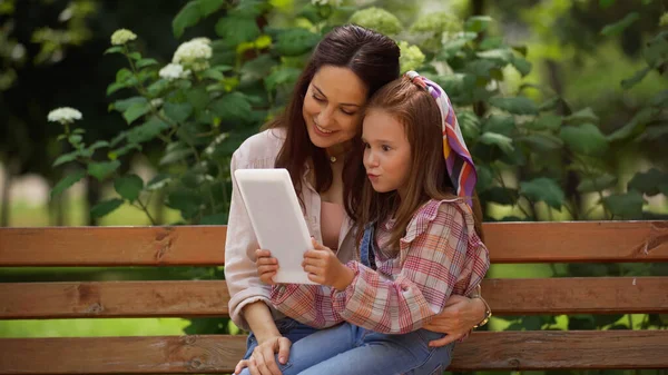 Smiling woman hugging preteen daughter with digital tablet on bench in park — Stock Photo