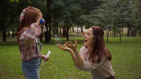 Positive woman standing near daughter blowing soap bubbles in park — Stock Photo