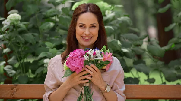 Cheerful woman holding bouquet and looking at camera on bench in park - foto de stock