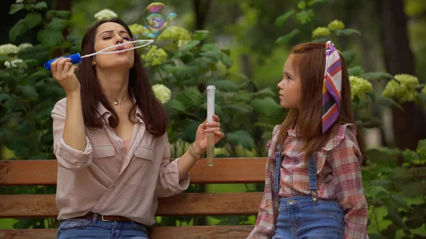Woman blowing soap bubbles near redhead daughter on bench in park — Stock Photo