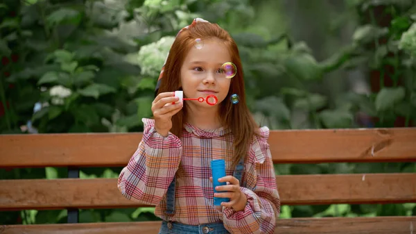 Red haired kid holding soap bubbles on bench in summer park — Stock Photo