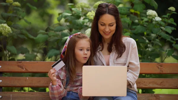 Smiling woman using laptop near near excited daughter with credit card on bench in park — Stock Photo