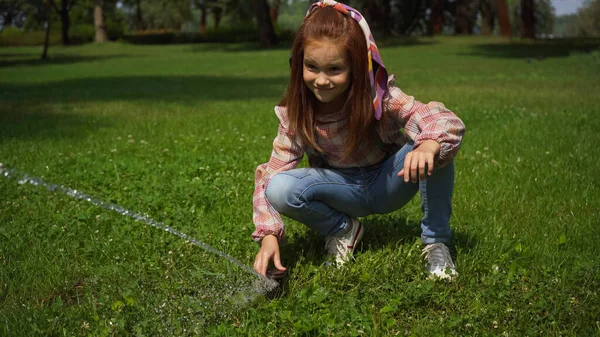 Red haired girl playing with automatic water sprayer in summer park — Fotografia de Stock