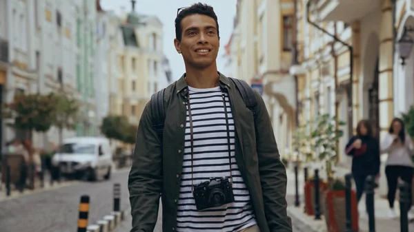Smiling bi-racial tourist with film camera walking on urban european street - foto de stock