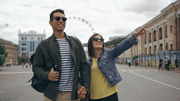 Smiling woman in sunglasses pointing with finger near bi-racial boyfriend on urban street — Stock Photo