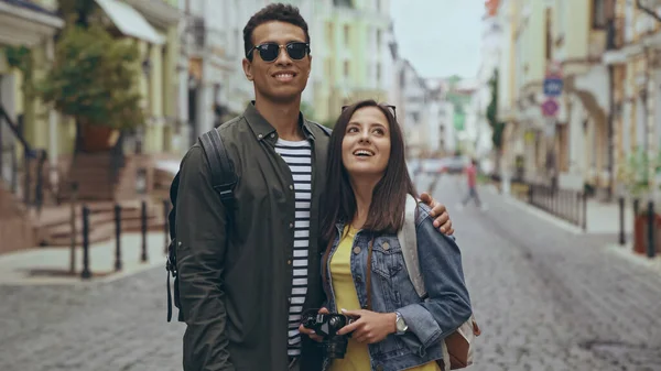 Smiling multiethnic tourists with backpack and retro camera standing on urban street — Stock Photo