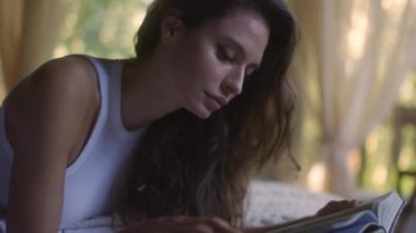 A young woman reads a book on the bed in the bedroom interior. Side view of female enjoying romantic novel lying in bed in the evening. A student read a textbook in the bedroom. Relax time with a book in comfort cozy atmosphere. 