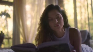 A young woman reads a book on the bed in the bedroom interior. Side view of female enjoying romantic novel lying in bed in the evening. A student read a textbook in the bedroom. Relax time with a book in comfort cozy atmosphere. 
