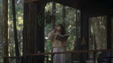 Kitap okuyan ve ormandaki sıcak bir kulübede sıcak çay içen genç bir kadın. Ormanın ortasındaki ahşap kulübe güneşli ahşap dağ kulübesi kırsal ev mimarisi..