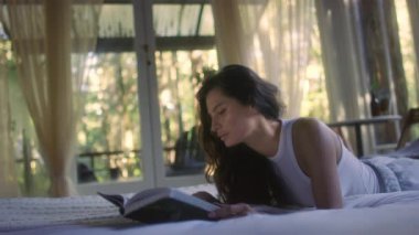 A young woman reads a book on the bed in the bedroom interior. Side view of female enjoying romantic novel lying in bed in the evening. A student read a textbook in the bedroom. Relax time with a book in comfort cozy atmosphere. 