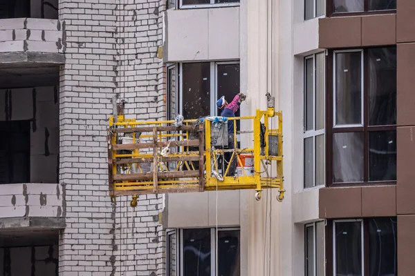 High-rise facade works. The builder in the yellow cradle. Work at height. Real estate market. Lifting mechanism.