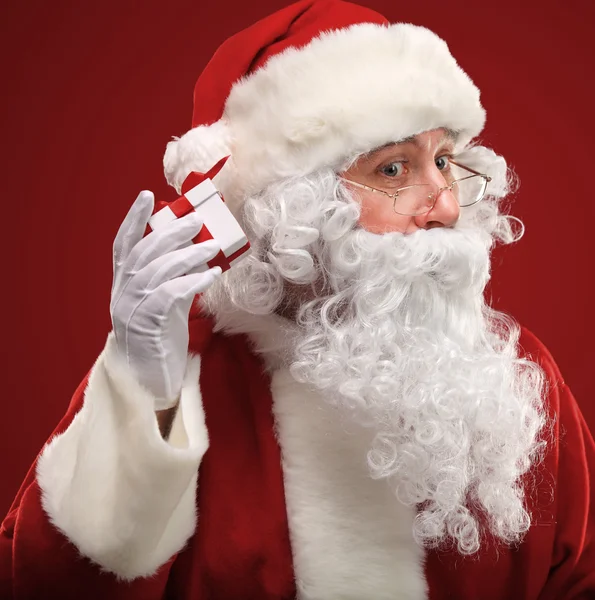 Photo du Père Noël tenant une boîte cadeau rouge près de son oreille — Photo