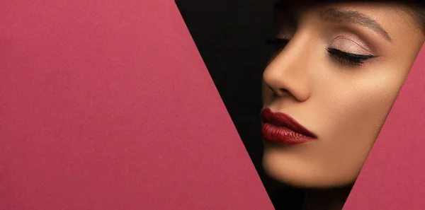 Ansiktet på en ung vacker flicka med en ljus make-up och med fylliga röda läppar kikar in i ett hål i rosa papper. Stockbild