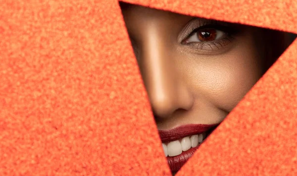 Das Gesicht eines jungen hübschen Mädchens mit leuchtendem Make-up und dicken roten Lippen guckt in ein Loch in glänzend rotem Papier. lizenzfreie Stockbilder