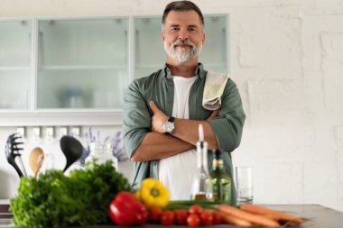 Mutfak masasında durmuş gülümseyen olgun bir adam kameraya bakıyor, sağlıklı yiyecekler hazırlıyor, taze sebze salatası yapıyor evde tek başına yemek pişirmenin keyfini çıkarıyor, portre yapıyor..