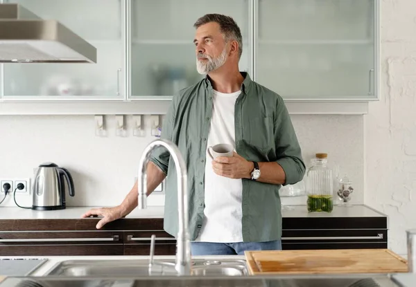 Heureux homme mûr buvant du café à la maison dans la cuisine, dégustant une boisson chaude le matin le week-end. — Photo