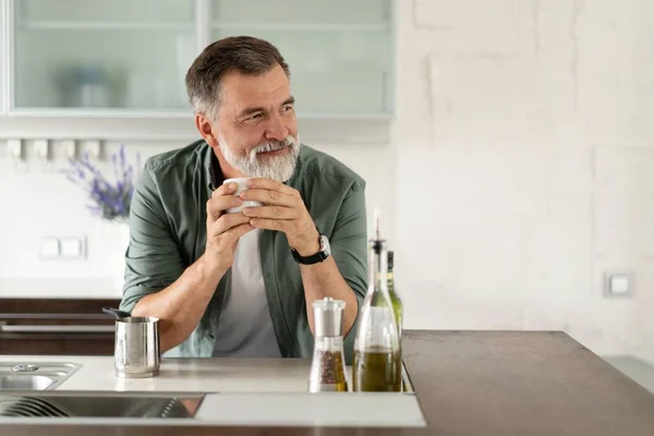 Szczęśliwy Dojrzały mężczyzna pije kawę w domu w kuchni, ciesząc się gorącym napojem rano w weekend. — Zdjęcie stockowe