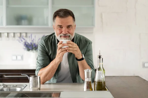 Boldog felnőtt férfi kávézik otthon a konyhában, élvezi a forró italt reggel hétvégén. — Stock Fotó