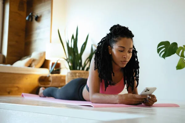 Jovem mulher esportiva depois de praticar ioga, quebrar fazendo exercício, relaxar no tapete de ioga, mensagens de texto no smartphone — Fotografia de Stock