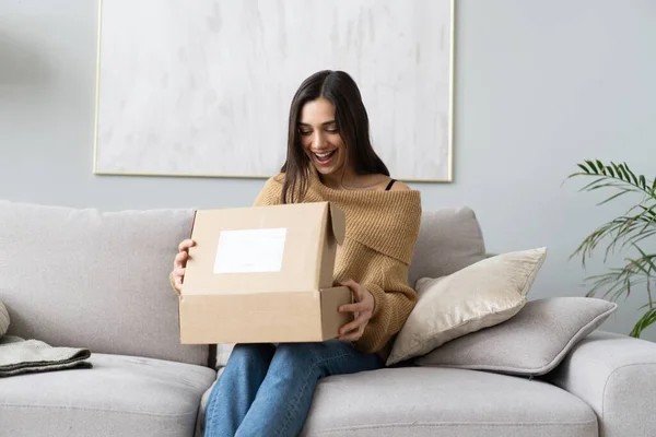 Mujer joven y feliz sentarse en la sala de sofá desempacar caja de cartón de compra de bienes en Internet. compras en línea, concepto de entrega — Foto de Stock