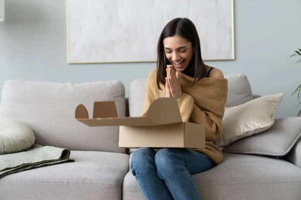 Wanita muda yang bahagia duduk di ruang sofa membongkar kotak kardus membeli barang-barang di Internet. belanja online, konsep pengiriman — Stok Foto