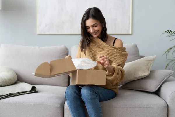 Mujer joven y feliz sentarse en la sala de sofá desempacar caja de cartón de compra de bienes en Internet. compras en línea, concepto de entrega —  Fotos de Stock