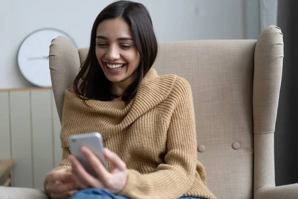 Jovencita sonriente sosteniendo el teléfono inteligente en las manos charlando en las redes sociales, utilizando aplicaciones móviles en casa. — Foto de Stock