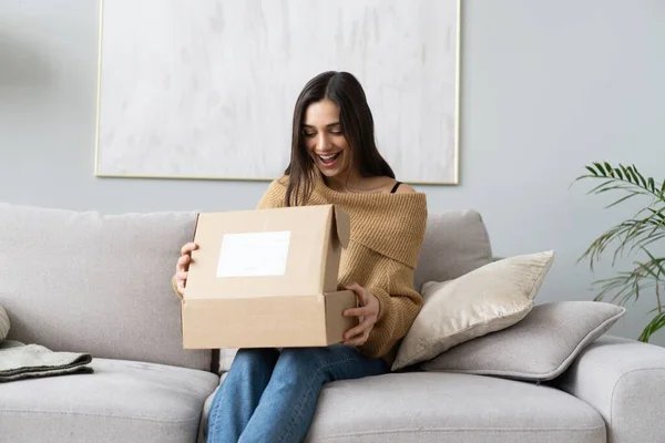 Mujer joven y feliz sentarse en la sala de sofá desempacar caja de cartón de compra de bienes en Internet. compras en línea, concepto de entrega Fotos de stock libres de derechos