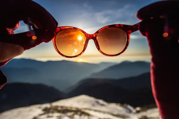Manos femeninas sosteniendo gafas de sol a través de las cuales se puede ver una hermosa puesta de sol entre las montañas de colores azul y naranja. — Foto de Stock