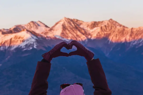 Frau macht Herzform mit den Händen über schneebedeckten Bergen in der Winterdämmerung — Stockfoto