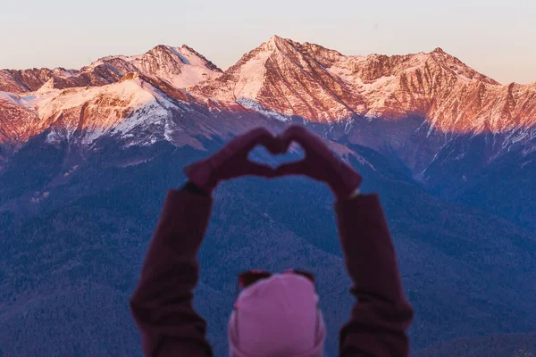 Frau macht Herzform mit den Händen über schneebedeckten Bergen in der Winterdämmerung — Stockfoto