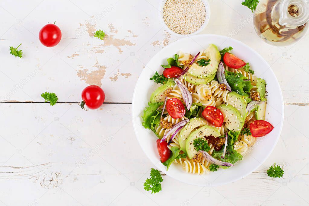 Fusilli pasta salad with avocado, tomatoes, fresh green lettuce, red onion and mustard dressing on white background. Vegetarian healthy lunch. Top view, flat lay