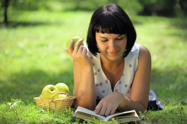 Young woman holding apple and reading book on grass — Stock Photo, Image