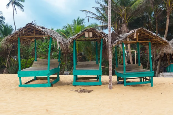 Drie lege turquoise zonnebank bedekt met palm tree bladeren aan zandstrand. — Stockfoto
