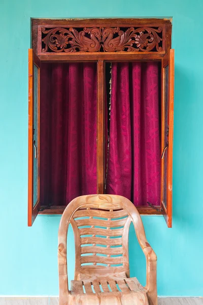 Plastikstuhl vor dem geöffneten Fenster mit Holzdekoration und Vorhängen aus Cyclamen. Stockbild