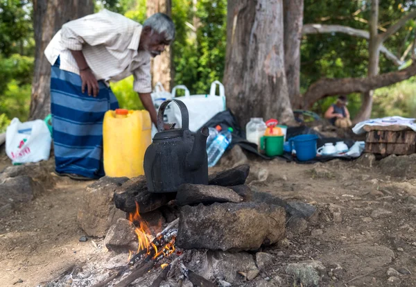 Homem local idoso fervendo água em uma panela no fogo do acampamento na floresta . — Fotografia de Stock