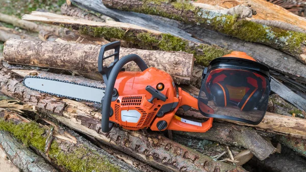 Portable power chain saw and safety helmet on heap of mossy wood logs. Orange chainsaw and hard hat with face guard and hearing protection. Occupational safety and health at work with sawing machine.