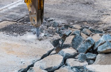 Toz, kıvılcım ve kırık beton parçalarıyla yıkım çekicinin yakın çekimi. Hidrolik kaya matkabının ayrıntıları eski yol yapılandırmasında çalışan kazıcı koluna monte edildi. İnşaat mühendisliğinde tamirat.