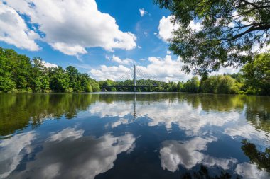 Shiny rippled water surface with reflection of white clouds on blue sky and green trees on a bank. Idyllic summer landscape with road cable-stayed bridge over Jordan pond in South Bohemian Tabor city.
