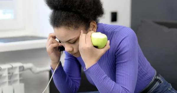 Afro-americana falando em um smartphone e comendo uma maçã. — Fotografia de Stock