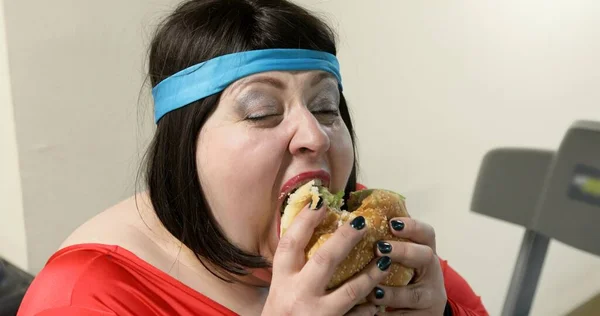Aç şişman kadın spor salonunda büyük bir hamburger yiyor.. 