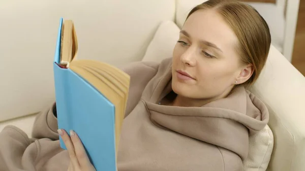 Roztomilá dívka sedí na gauči a čte fascinující knihu. — Stock fotografie