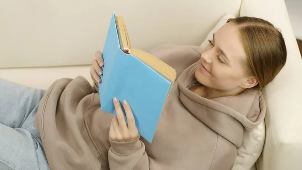 Een schattig meisje zit op de bank en leest een fascinerend boek. — Stockfoto