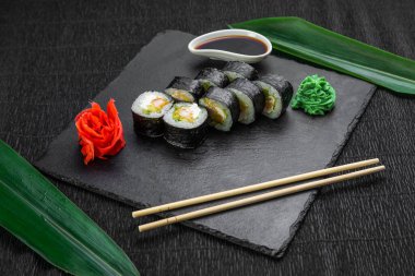 Suşi ruloları bambu yaprakları ve çubuklarla süslenmiş karanlık bir arka planda serili.