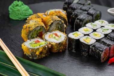 Suşi ruloları bambu yaprakları ve çubuklarla süslenmiş karanlık bir arka planda serili.