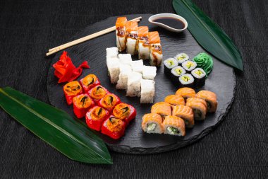 Suşi ruloları bambu yaprakları ve çubuklarla süslenmiş karanlık bir arka planda serili.