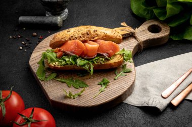 Somon balıklı sandviç ve roka. Koyu bir arka planda tahta bir tahtada servis ediliyor.