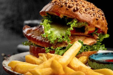 Biftek, marul ve peynirli hamburger. Koyu bir arka planda patates kızartması ve sos ile servis edilir.