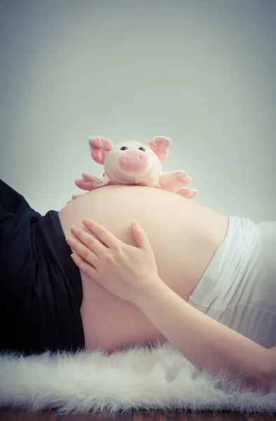 Pregnant belly lying on white fur with pink piggy — Stock Photo, Image